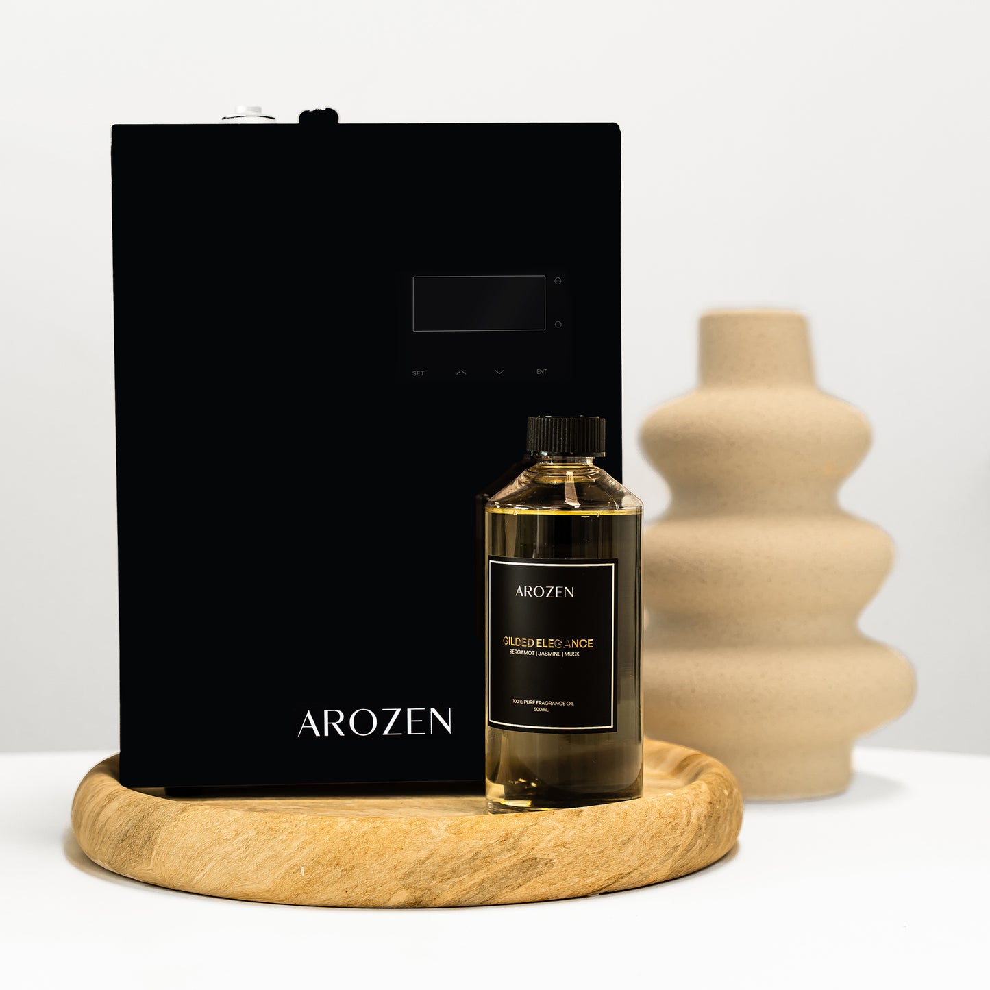 A black Arozen Vortex Ultra electric Scent Diffuser and a bottle of Arozen fragrance oil rest on a round wooden tray, with a beige abstract vase in the background against a white surface.