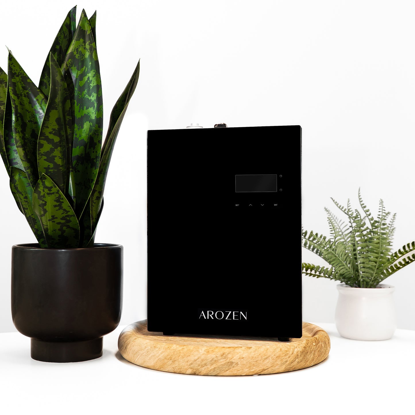 A black rectangular Arozen VORTEX ULTRA ELECTRIC SCENT DIFFUSER sits on a round wooden tray between a tall leafy plant in a black pot and a small fern in a white pot, ready to disperse fragrance oils with cold-air diffusion against a white background.