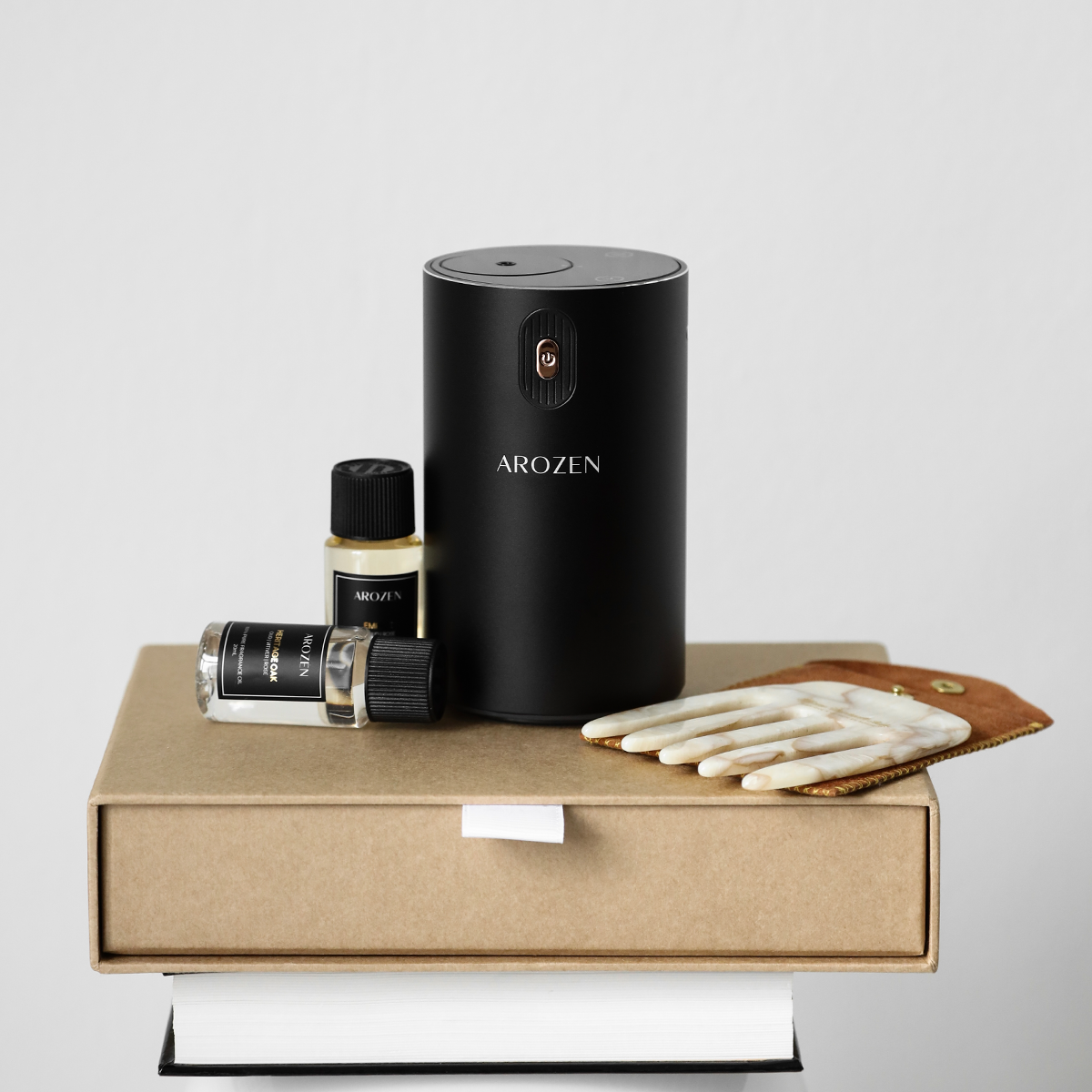 A sleek black cylindrical EON MINI ELECTRIC SCENT DIFFUSER by Arozen stands on a box next to two fragrance oil bottles —all arranged in minimalist style.