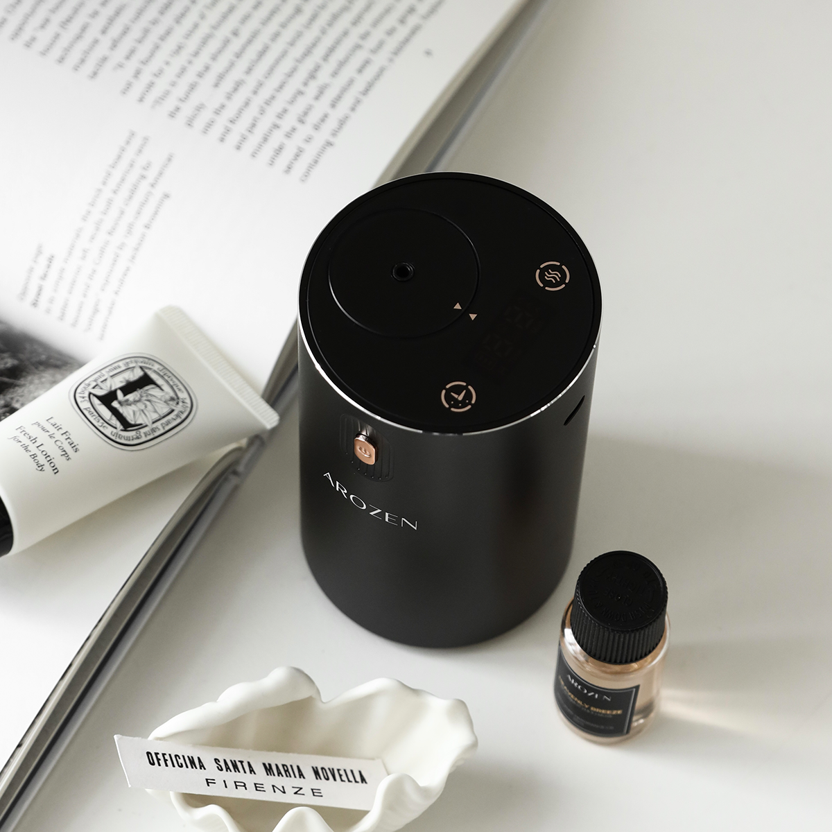 A sleek black cylindrical EON MINI ELECTRIC SCENT DIFFUSER by Arozen in a top down view stands on table next to a fragrance oil bottles and a book — all arranged in minimalist style.