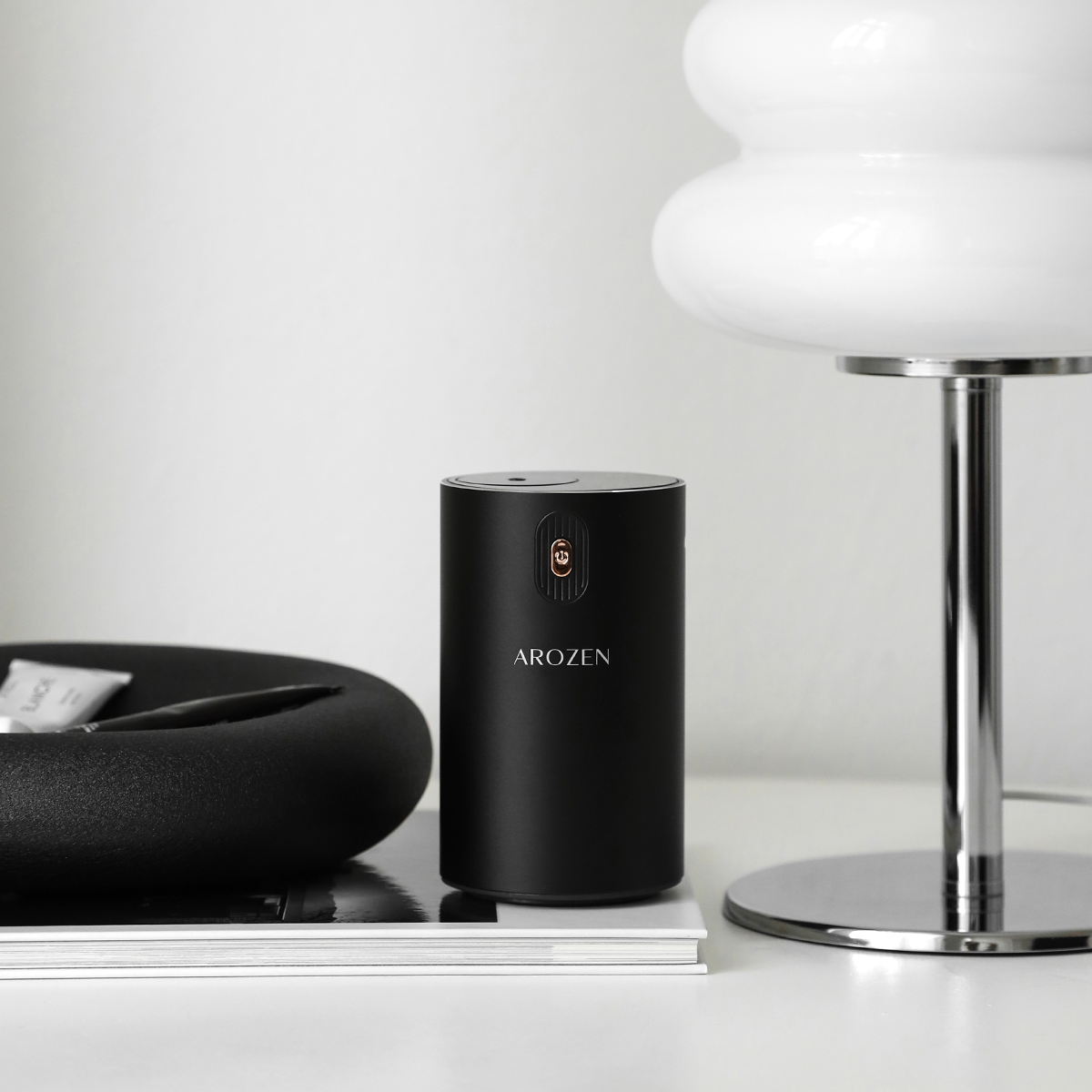 A sleek black cylindrical EON MINI ELECTRIC SCENT DIFFUSER by Arozen stands on a book next to a lamp—all arranged in minimalist style.
