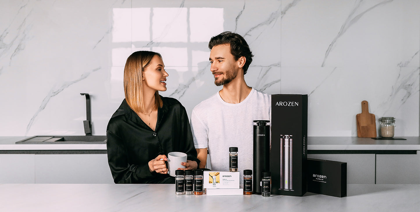 A woman and a man stand in a modern kitchen, smiling at each other. Various Arozin-branded electric scent diffuser products are displayed on the countertop in front of them. The woman holds a white mug.