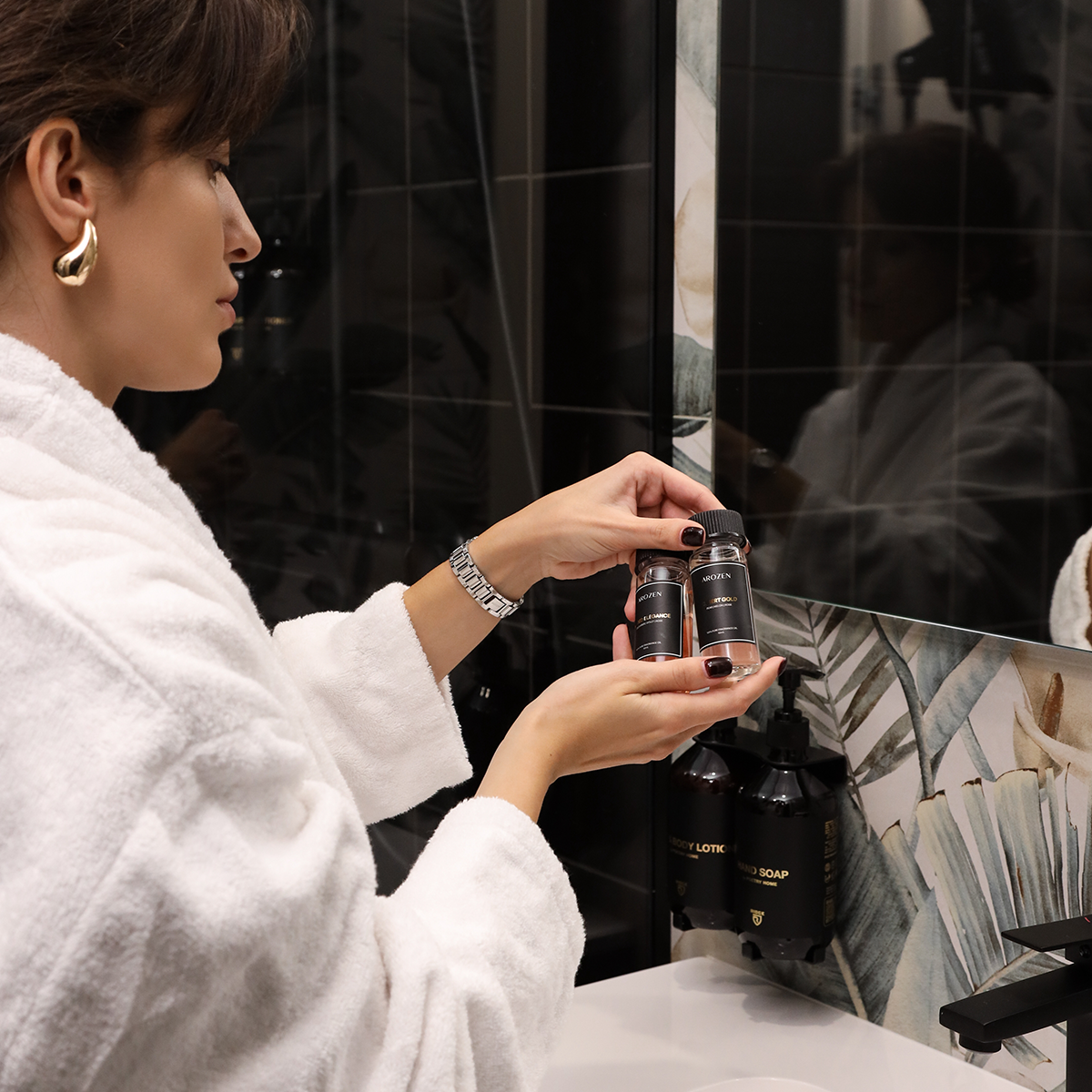A woman in a white bathrobe stands in a bathroom, holding and examining two small black bottles of Arozen electric scent diffuser fragrance oil.